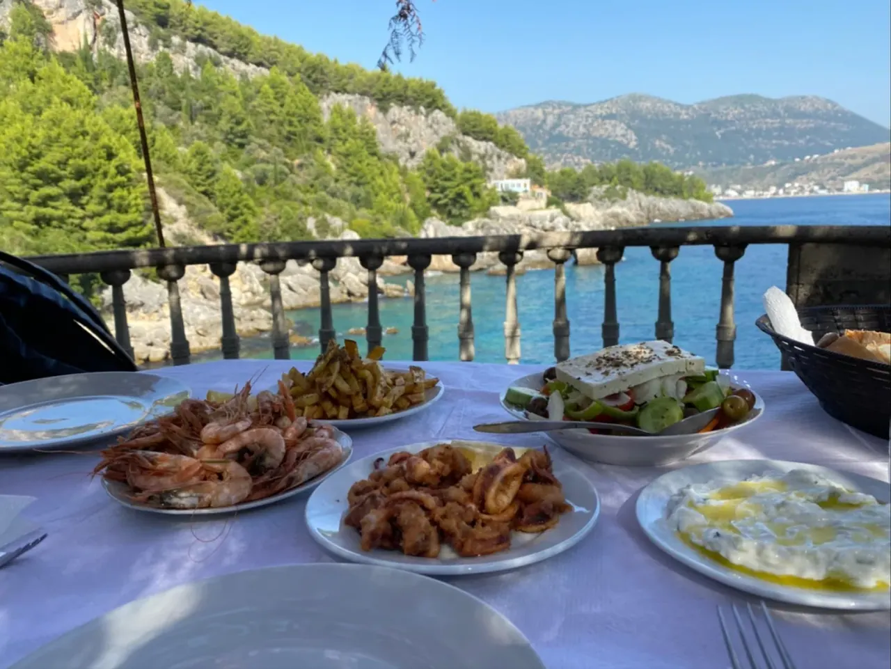 Table of fresh seafood and Greek salad with a view over a turquoise cove at Kiço Meniko in Himara