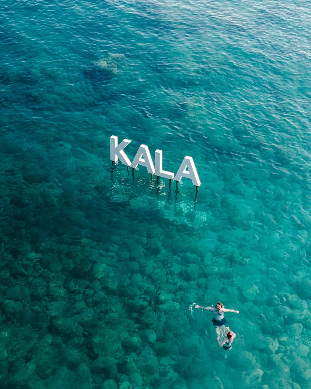 Aerial view of the iconic KALA letters standing in the turquoise shallows off Dhermi beach, with swimmers floating nearby — the Albanian Riviera setting of Kala Festival
