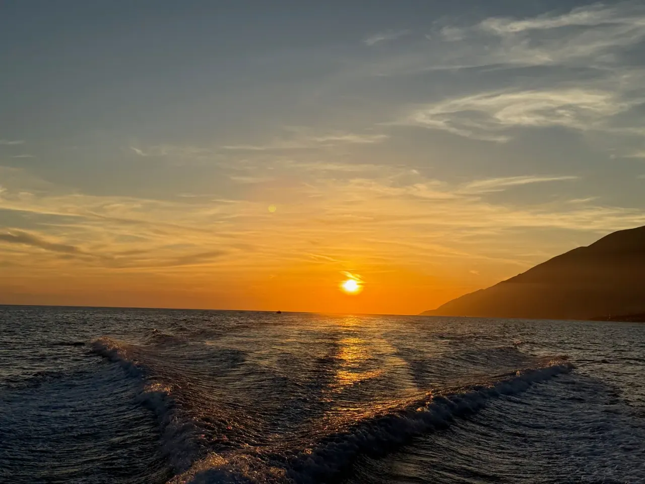 Golden-hour sun setting over the Ionian Sea behind the Ceraunian coastline, seen from a boat's wake off Himara